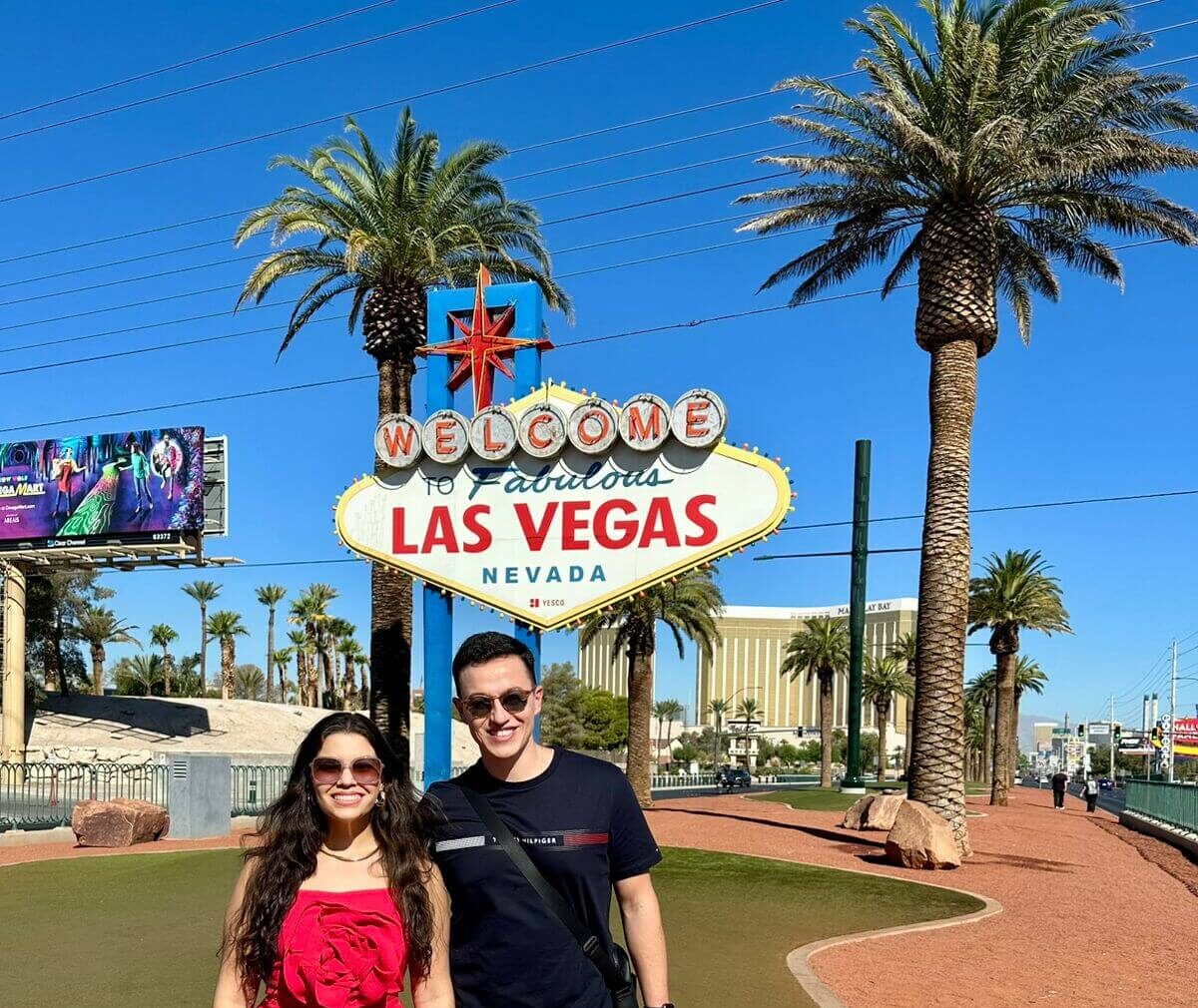 Casal posando ao lado do famoso letreiro de "Welcome to Fabulous Las Vegas", com céu azul e clima desértico ao fundo.