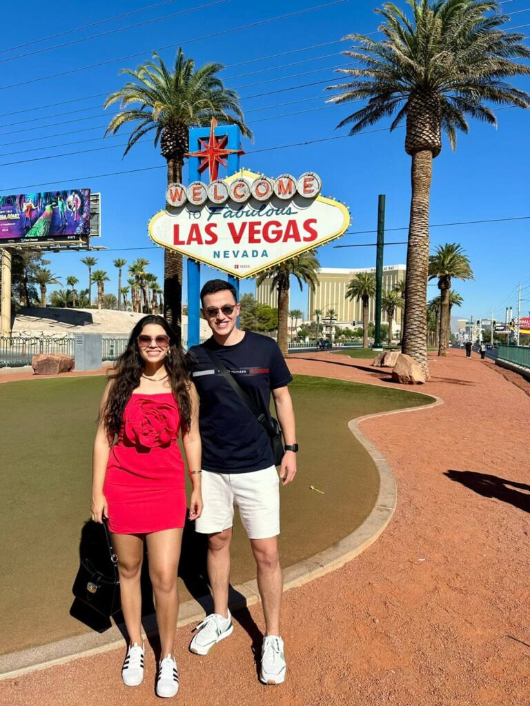 Casal posando ao lado do letreiro Welcome to Fabulous Las Vegas com céu azul ao fundo