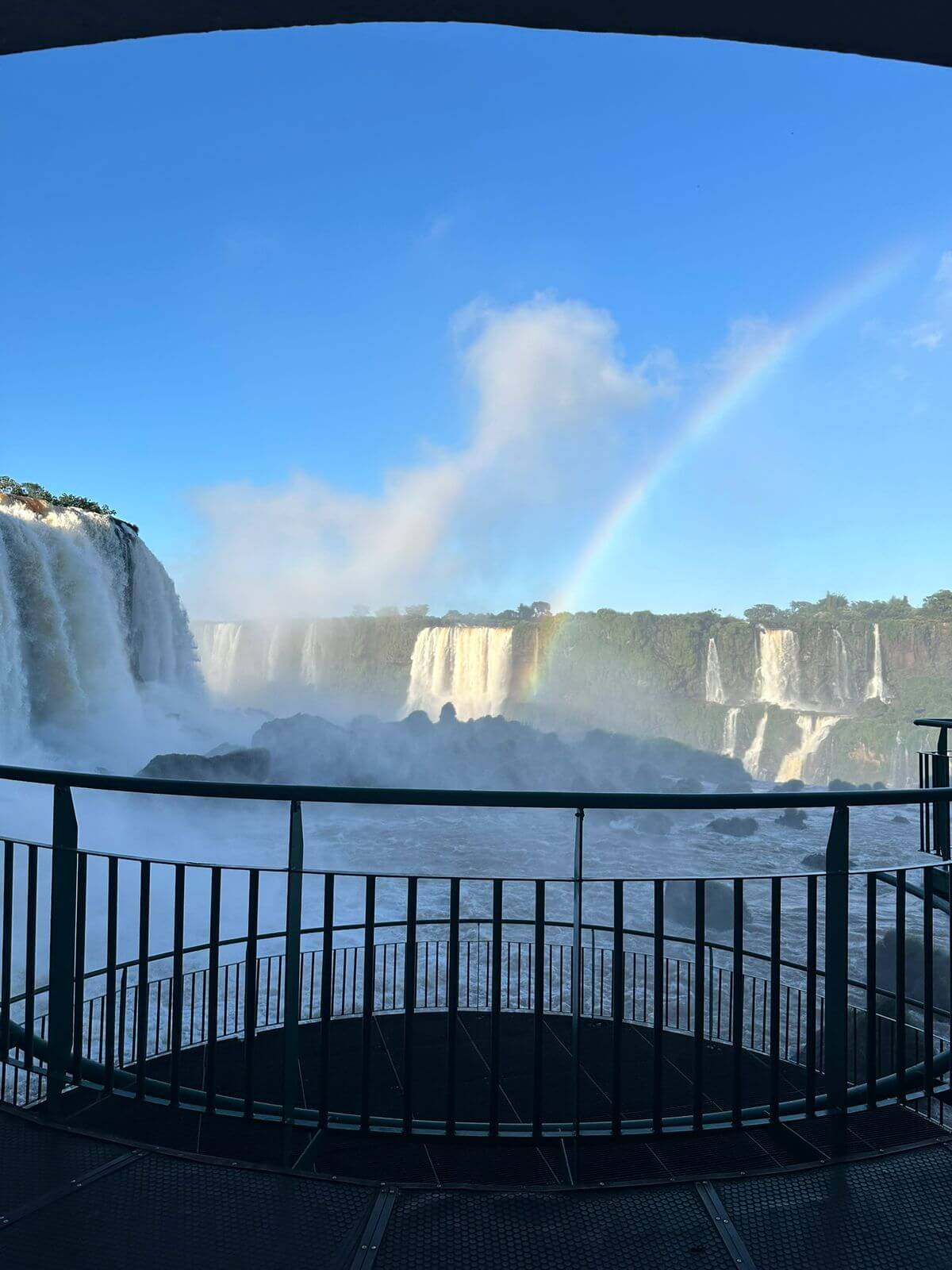 Vista das Cataratas do Iguaçu a partir da passarela, com arco-íris formado pela neblina em dia ensolarado.