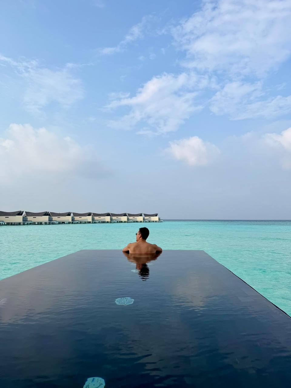Casal na piscina privativa de borda infinita da vila sobre as águas do Kuredhivaru Resort, Maldivas, com a lagoa cristalina e outras vilas ao fundo
