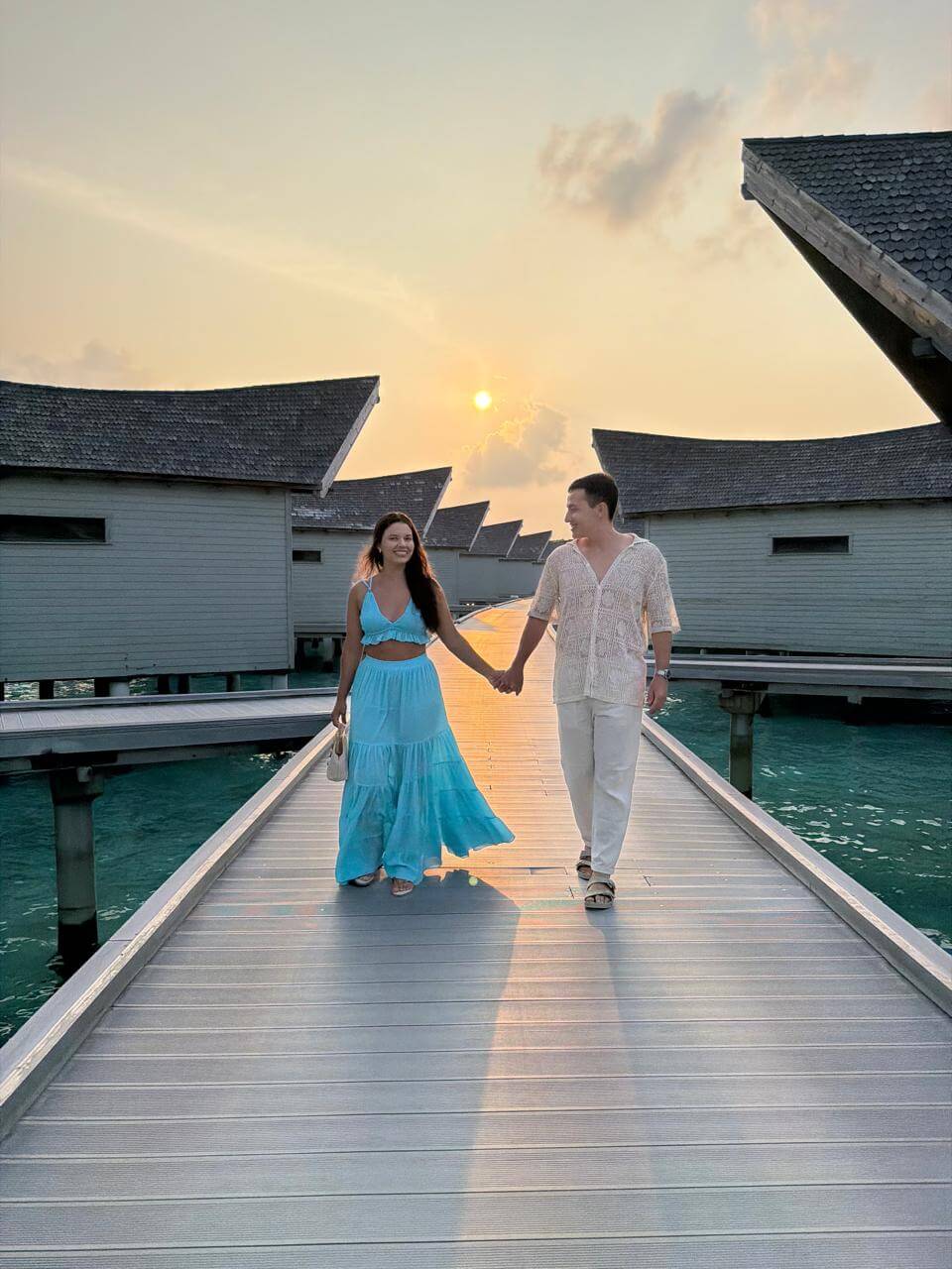 Casal caminhando de mãos dadas no deck entre as vilas sobre as águas do Kuredhivaru ao pôr do sol nas Maldivas