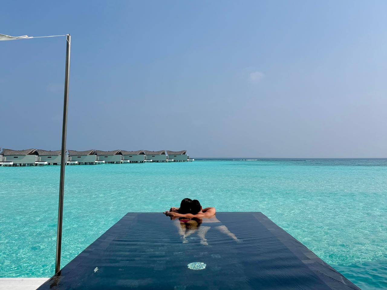 Casal na piscina privativa de borda infinita da vila sobre as águas do Kuredhivaru Resort, Maldivas, com a lagoa cristalina e outras vilas ao fundo