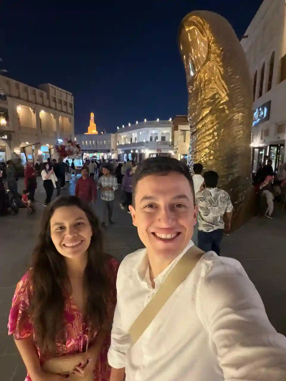 Casal sorrindo de noite no Souq Waqif em Doha, com a escultura do dedo dourado ao fundo