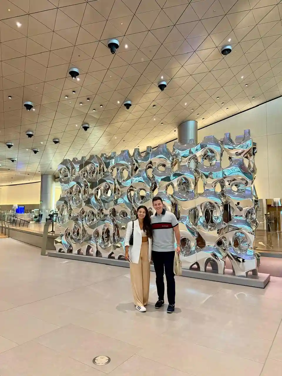 Casal em frente ao painel de esculturas metálicas dentro da Al Mourjan Business Lounge South no aeroporto de Doha