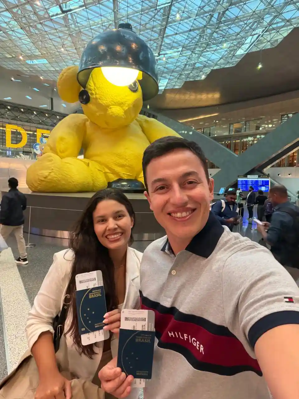 Casal com passaportes em frente ao urso amarelo icônico do Aeroporto Internacional Hamad de Doha