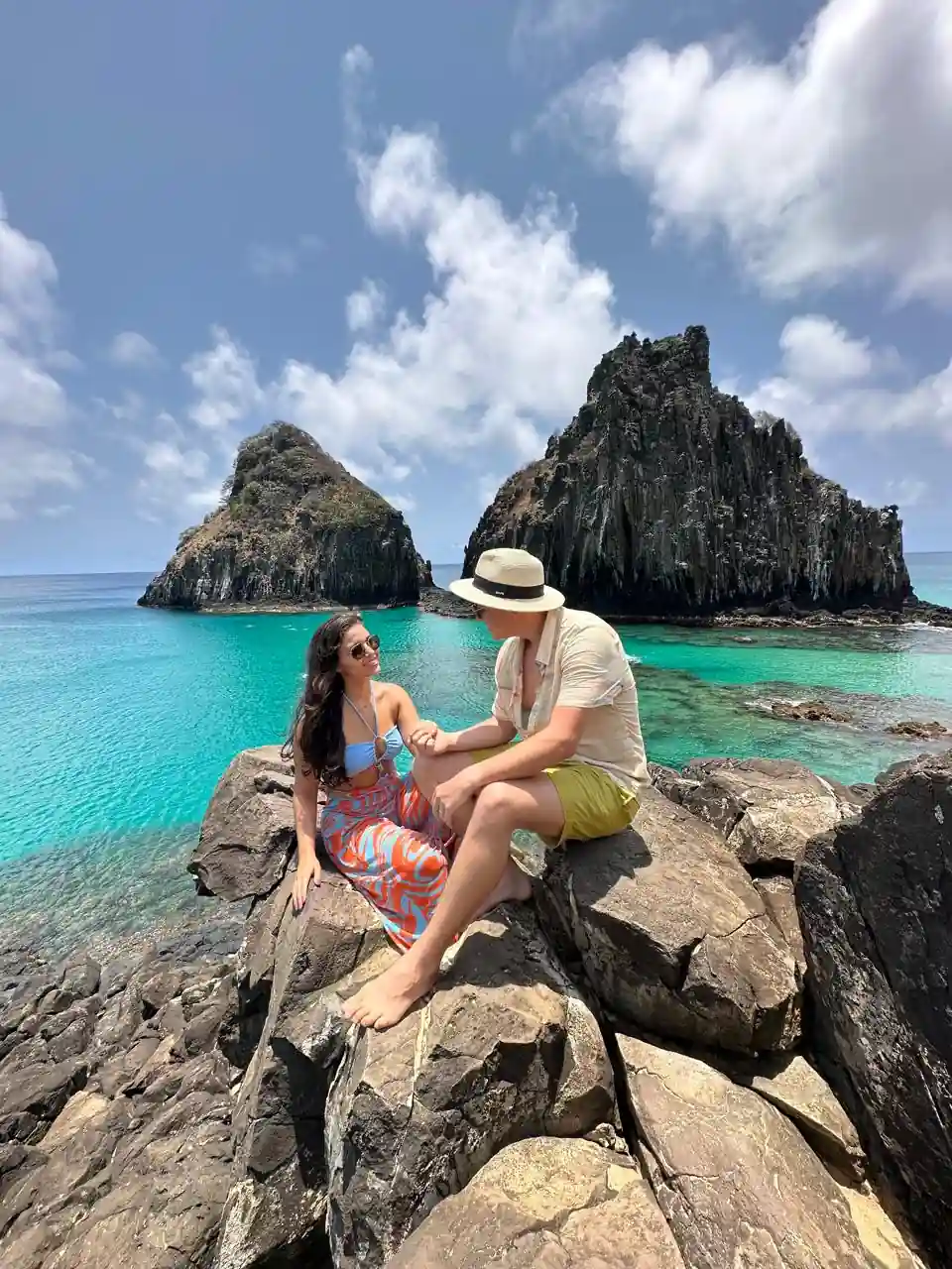 Casal sentado nas pedras em frente ao Morro Dois Irmãos em Fernando de Noronha