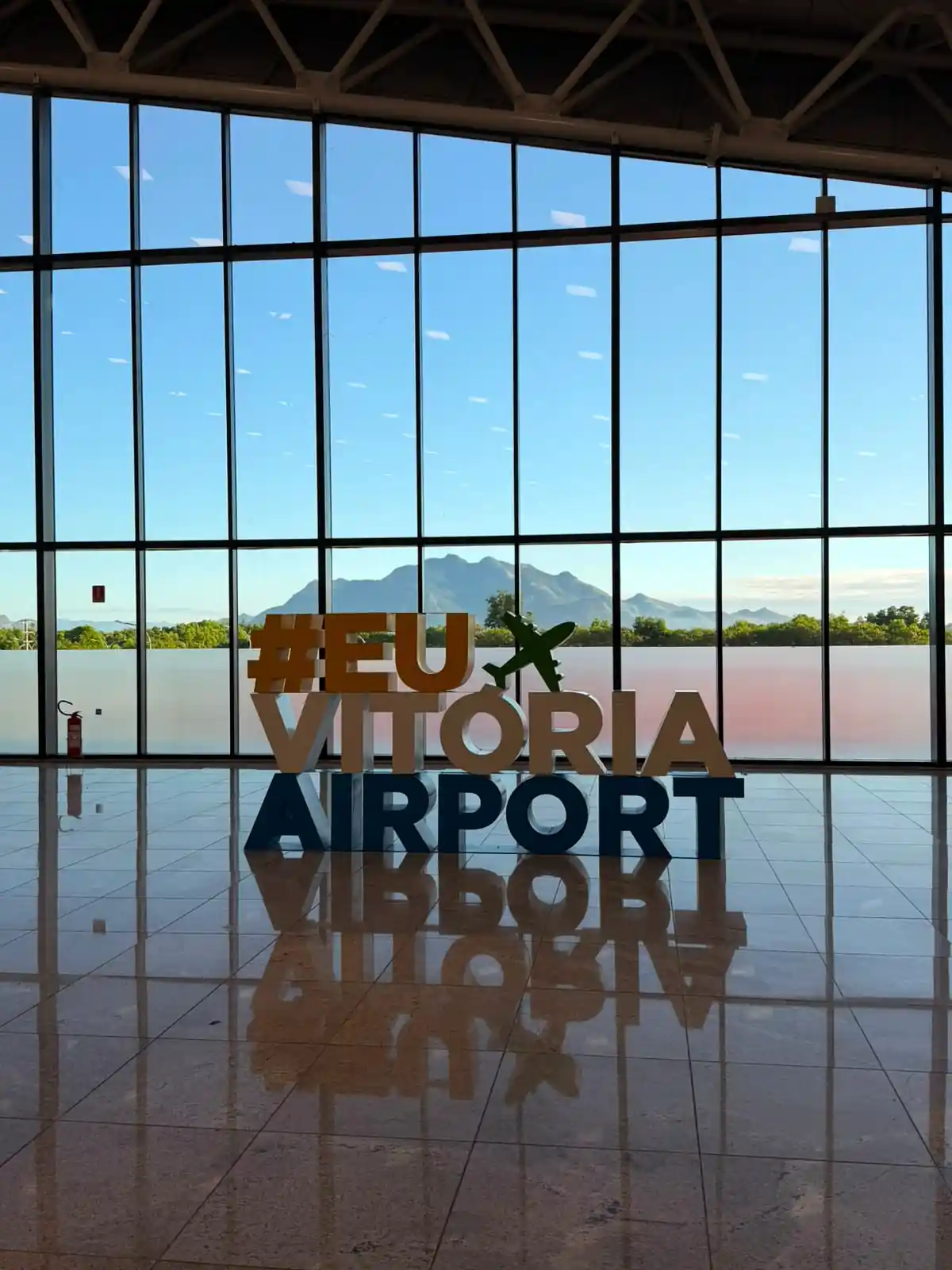 Letreiro #EuVitoriaAirport no terminal do Aeroporto de Vitória, com vista para as montanhas ao fundo